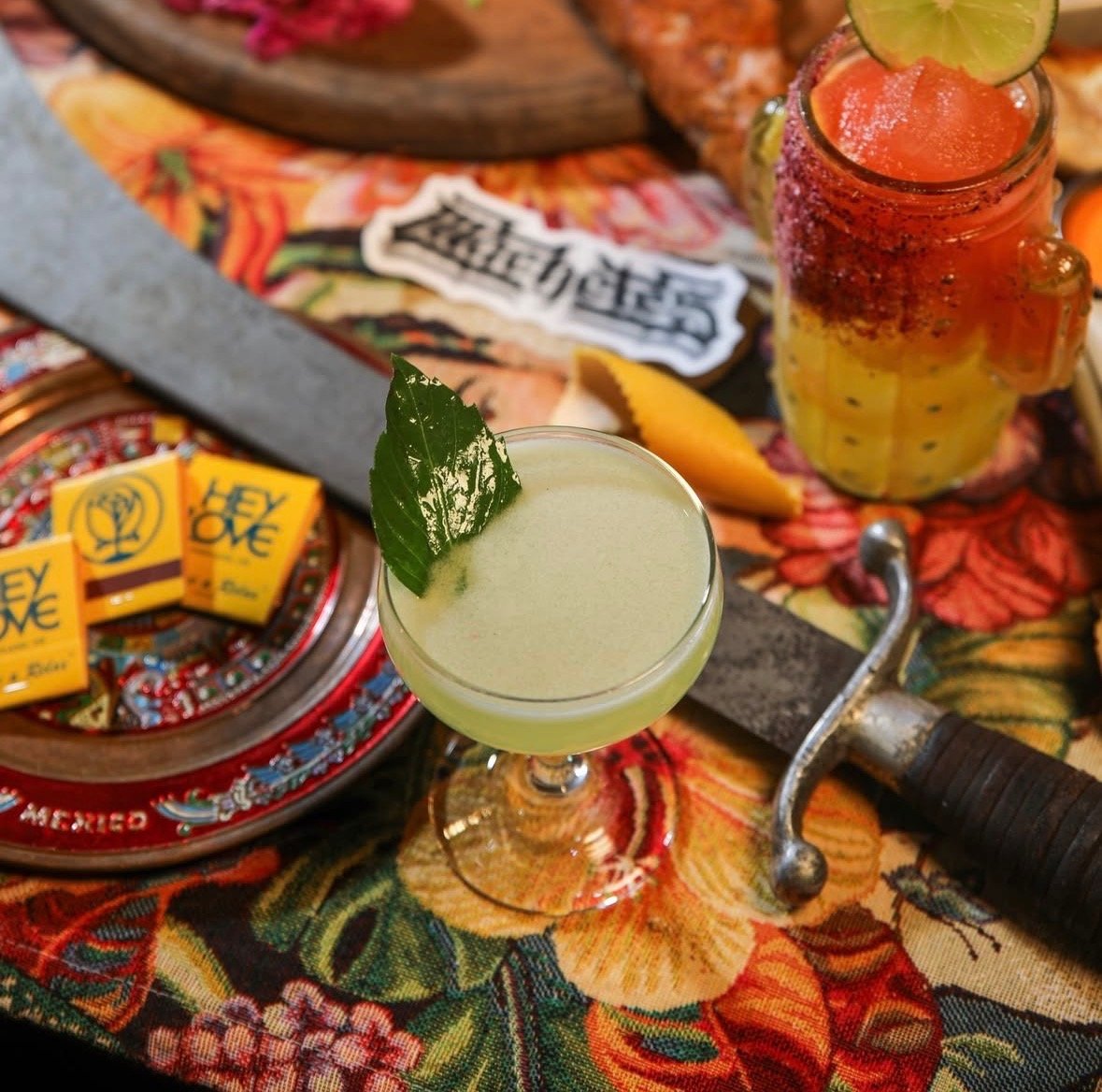 A bar setup with two cocktails, one green with a leaf garnish and one red with fruit slices, surrounded by a bag of tequila, lemon, a razor, and chili powder on a floral tablecloth.