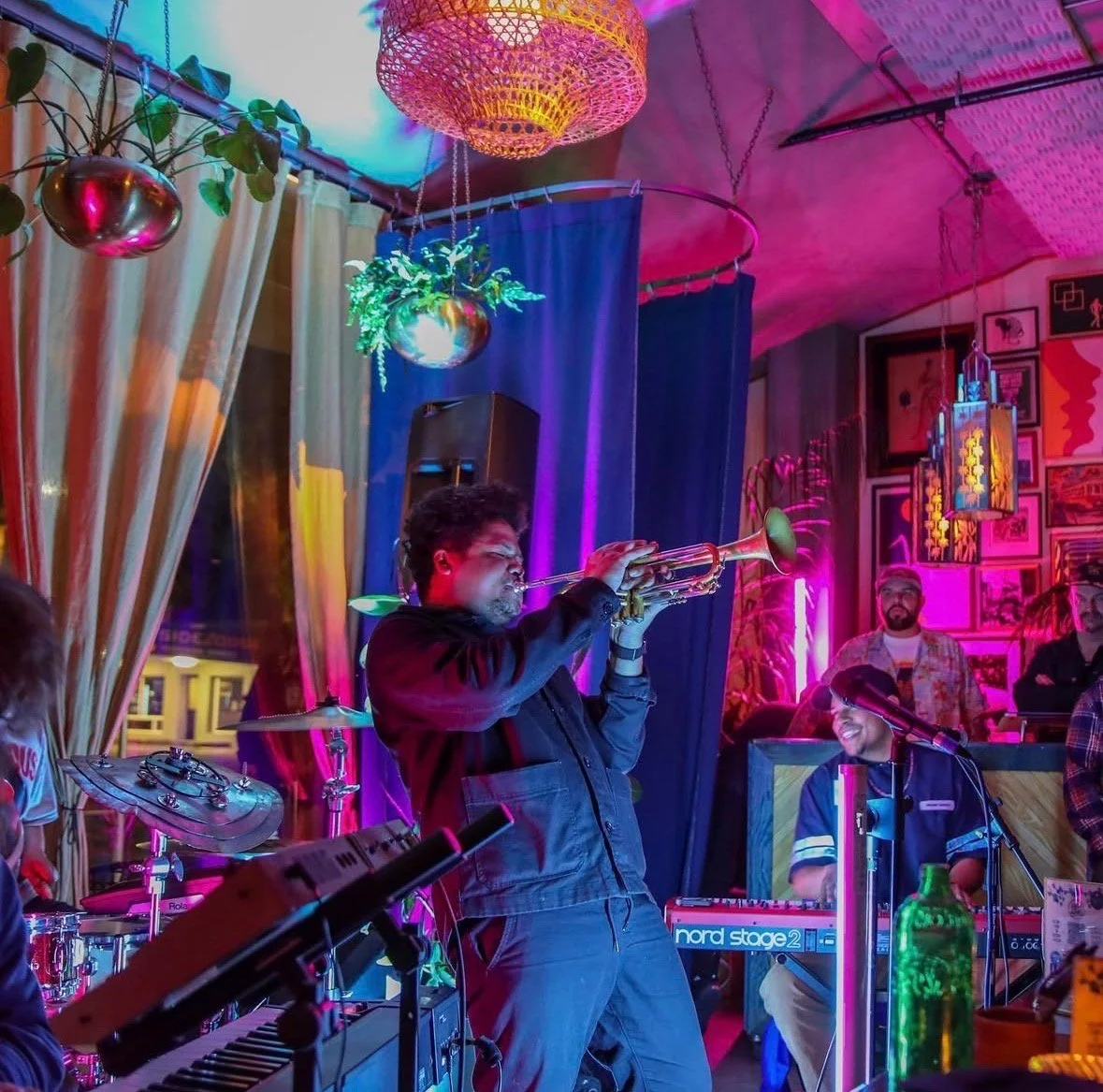 A man playing the trumpet on stage at a lively, colorful indoor music venue, surrounded by keyboard, drums, and other band members with vibrant lighting and wall art.