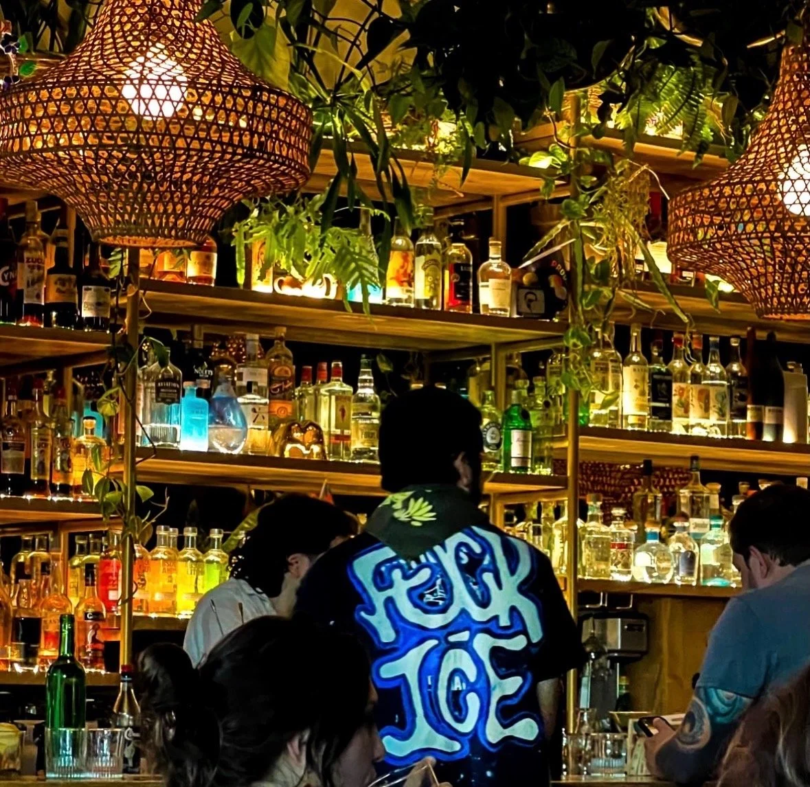 A bartender behind a well-stocked bar with bottles of liquor, surrounded by patrons, in a warm, ambient setting with hanging woven lamps and green foliage.