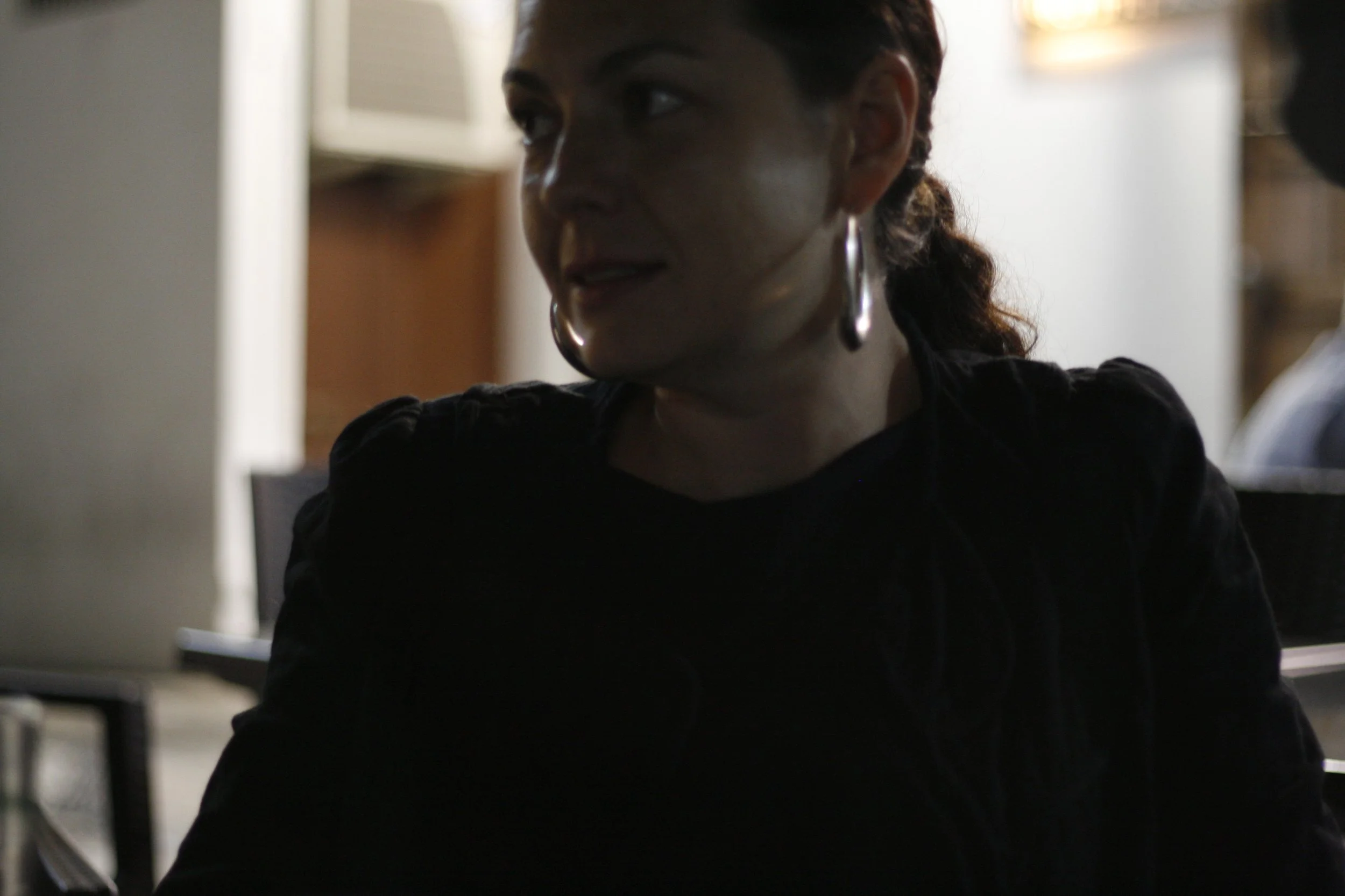 Retrato de una mujer con pendientes grandes, cabello rizado recogido y vestido negro, en un ambiente interior oscuro y con luz suave.