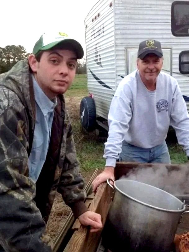 Two men standing beside a steaming pot outdoors near a camper trailer. One man is wearing a camouflage jacket and green cap, the other in a gray sweatshirt and black cap with a John Deere logo.