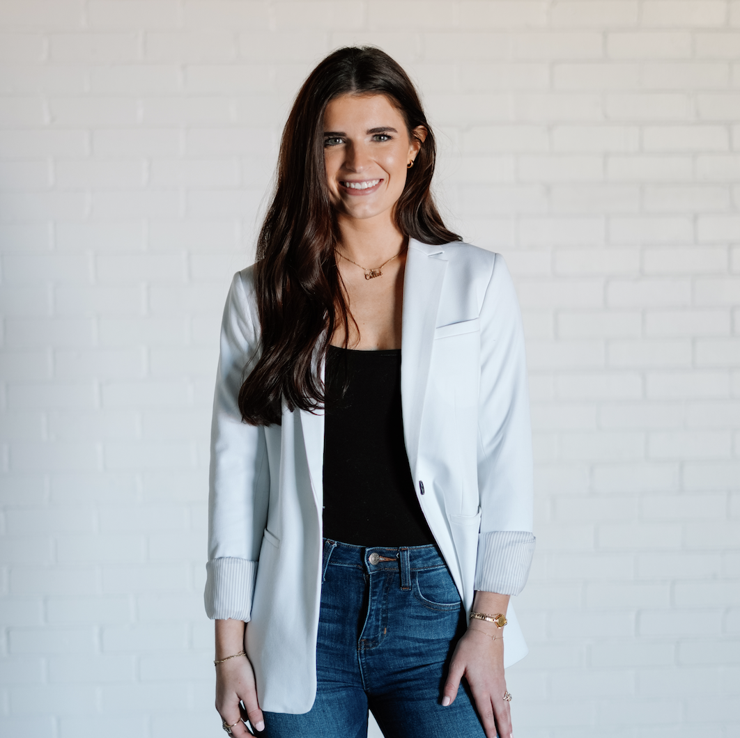 A woman with long dark hair wearing a white blazer, black top, and blue jeans standing against a white brick wall, smiling at the camera.