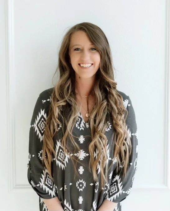 A young woman with long wavy hair wearing a black and white patterned blouse, smiling while standing against a light-colored wall.