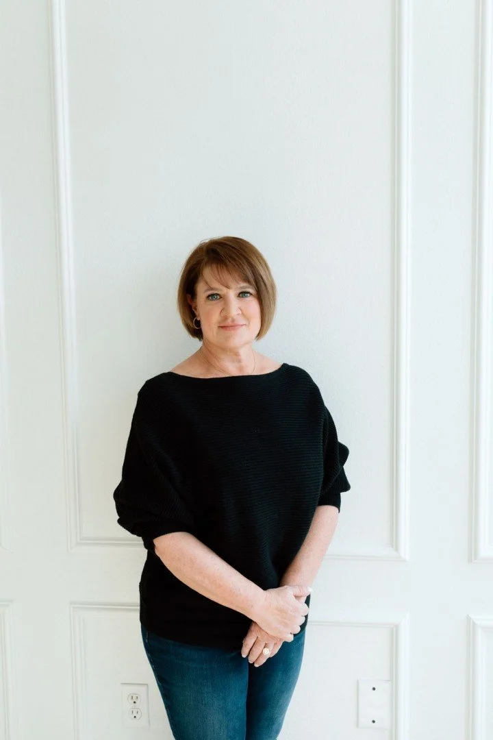 A woman with short brown hair standing against a white wall with decorative molding, wearing a black sweater and blue jeans.