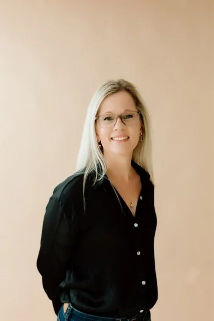 A woman with long blonde hair, glasses, and earrings, smiling and wearing a black button-up shirt, standing against a plain light background.