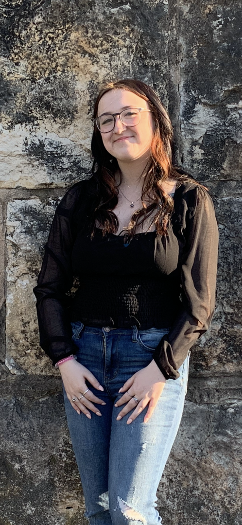 A young woman with glasses and long brown hair standing in front of a textured stone wall, smiling softly, dressed in a black sheer long-sleeve top and ripped jeans.