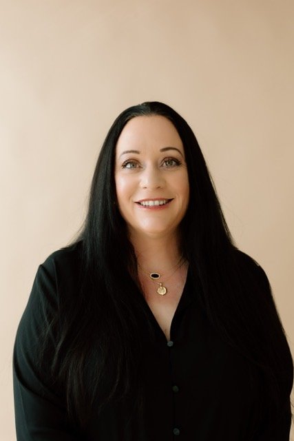 Portrait of a woman with long black hair, wearing a black top, and a gold necklace, smiling against a plain beige background.