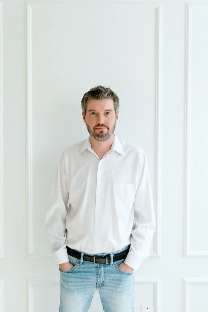 A man with dark hair and a beard stands against a white paneled wall, wearing a white button-down shirt and light blue jeans with his hands in his pockets.