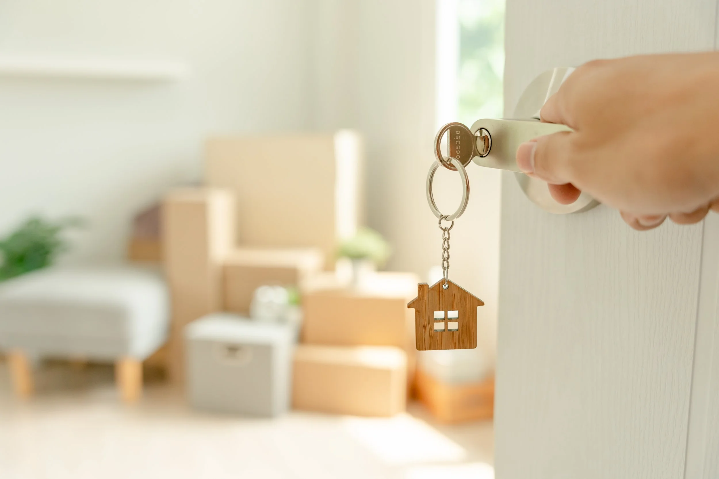 Close-up of a hand inserting a key into a lock on a door, with a small wooden house-shaped keychain hanging from it, in a brightly lit room.