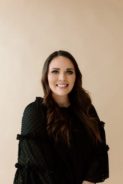 Young woman with long brown hair smiling, wearing a black long-sleeve top, in front of a beige background.