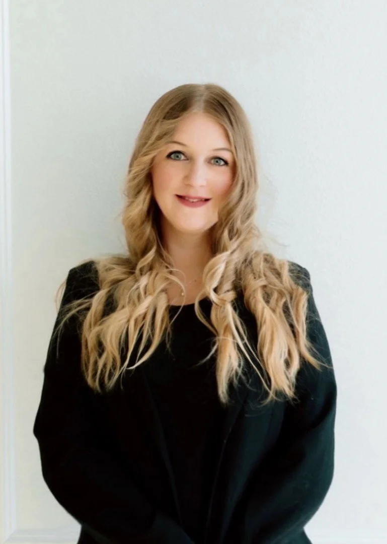 A young woman with long, wavy blonde hair and blue eyes, smiling, standing against a plain white wall, wearing a black outfit.