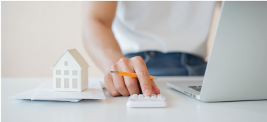 Person using a calculator at a desk with a small house model and a laptop.