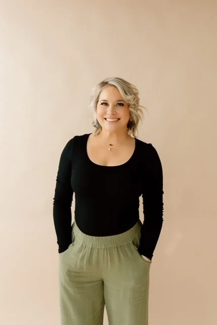 A woman with shoulder-length blonde hair smiling, wearing a black long-sleeve top and light green pants, standing against a light beige background.