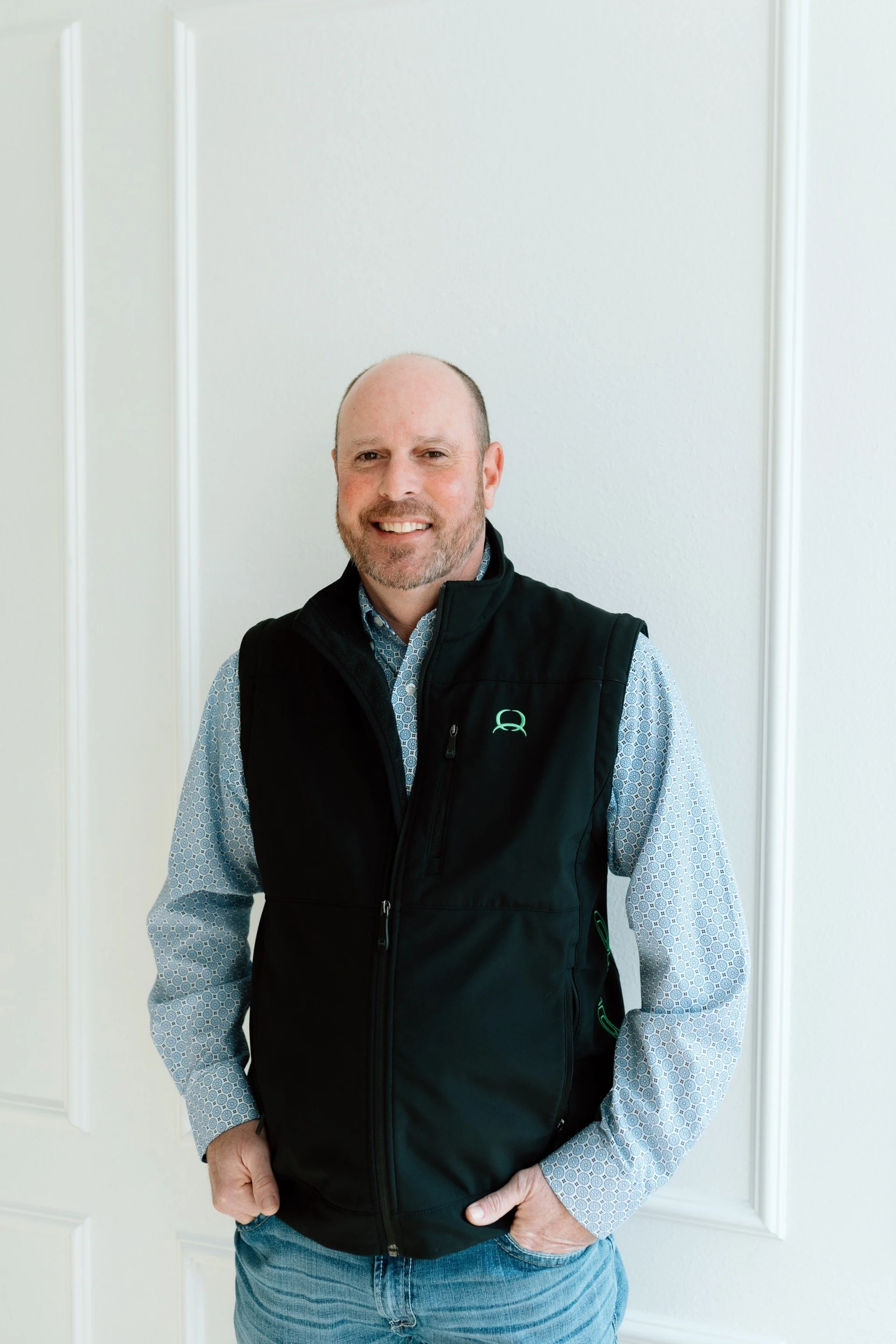 A middle-aged man with a beard and short hair, smiling, wearing a patterned button-up shirt and a black vest with a logo, standing in front of a plain white wall.