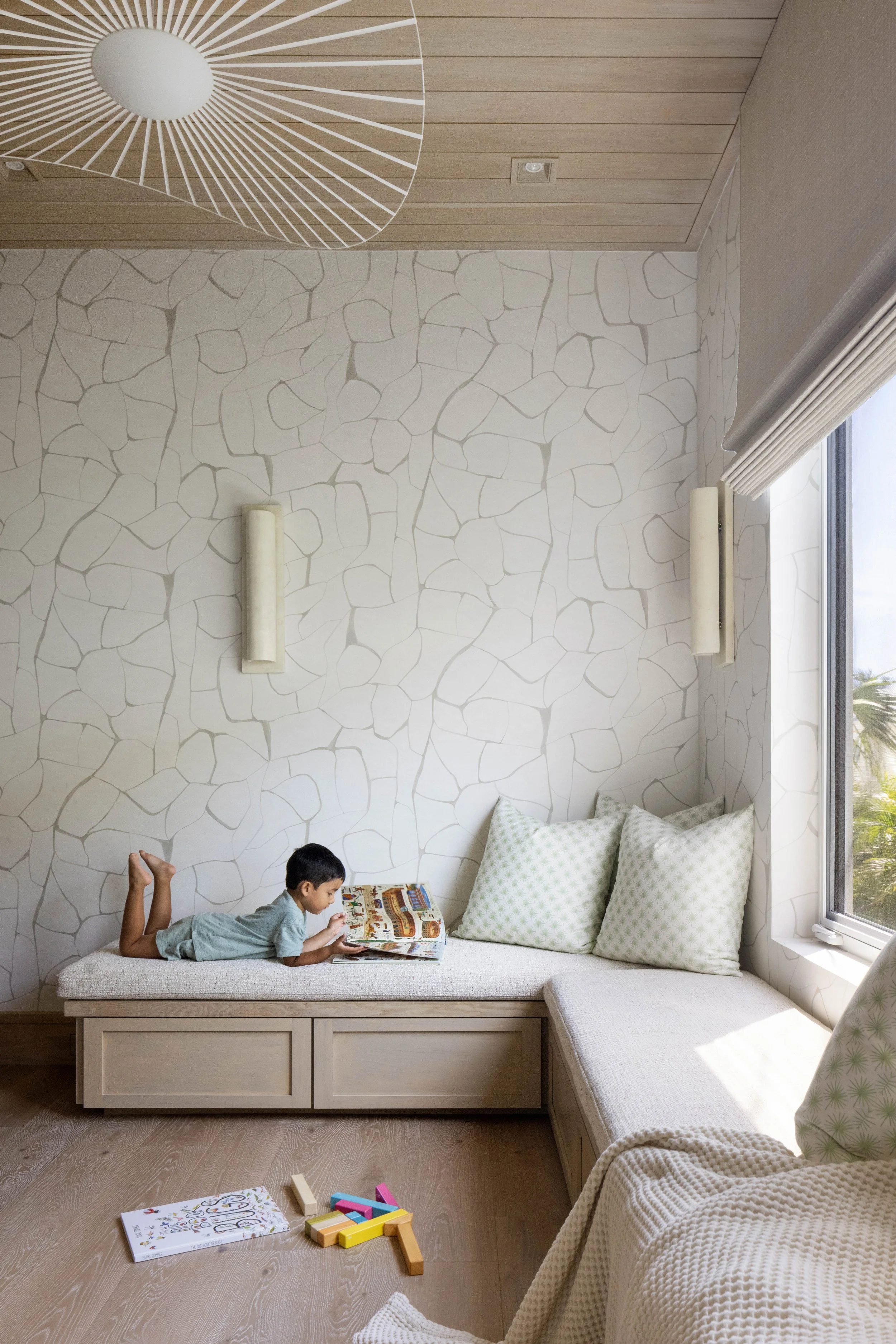 Young boy lying on a bed reading a book in a cozy room with patterned wallpaper, large pillows, and a window with blinds.