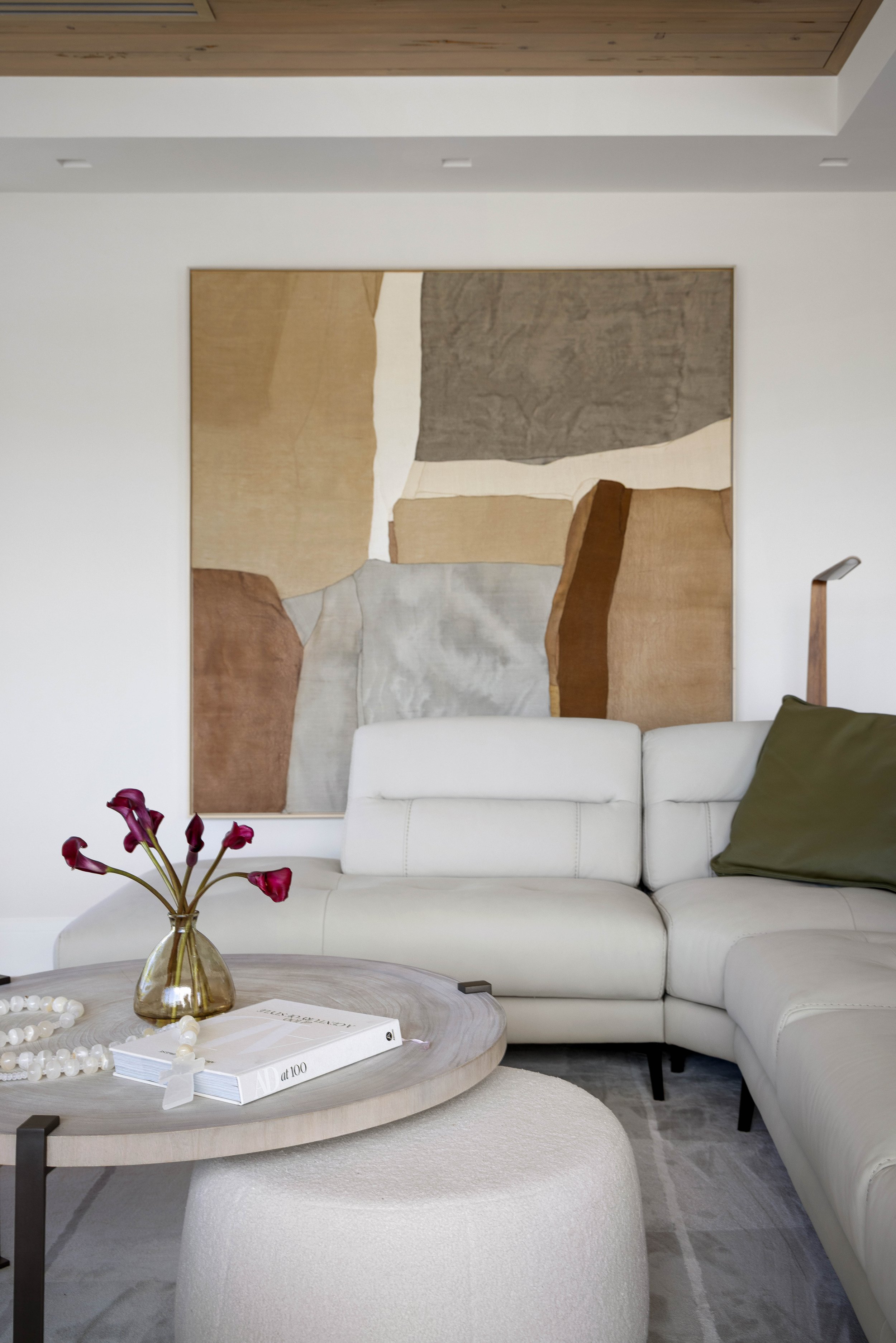 Living room with white sectional sofa, green pillow, round wooden coffee table with a vase of pink flowers, a book, and a large abstract artwork hanging on the wall background.