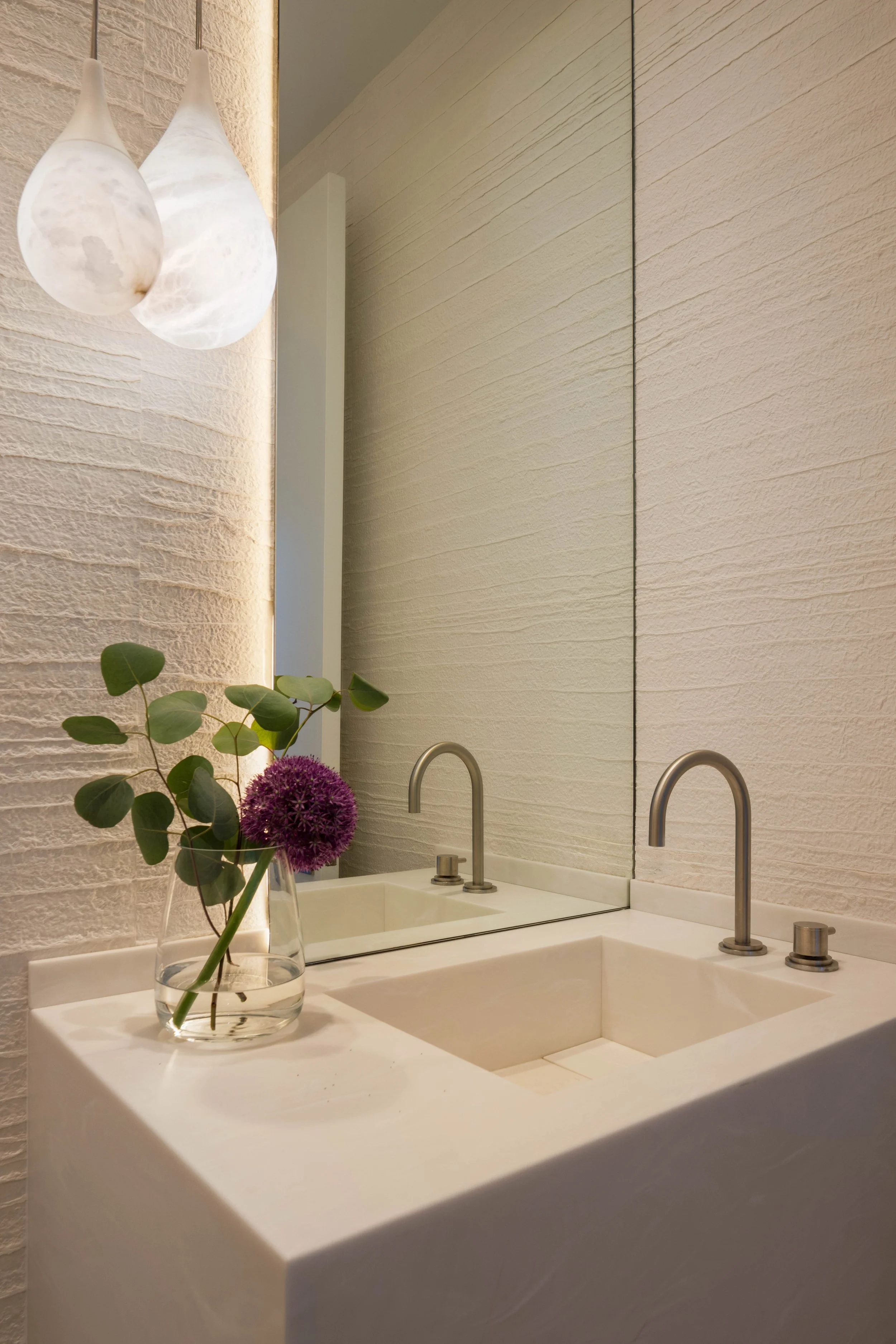 A bathroom vanity with a white countertop, a built-in sink, and two modern stainless steel faucets. A glass vase with green leaves and purple flowers is on the countertop. A large mirror and textured beige wall panels are in the background, illuminated by soft lighting.