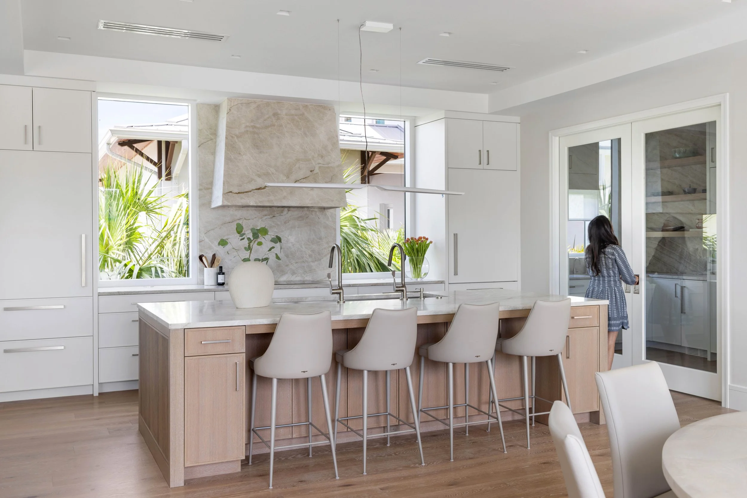 Modern kitchen with white cabinets, a large island with light wood finish, and a woman in a patterned dress standing near the sink. Large windows overlook greenery outside.
