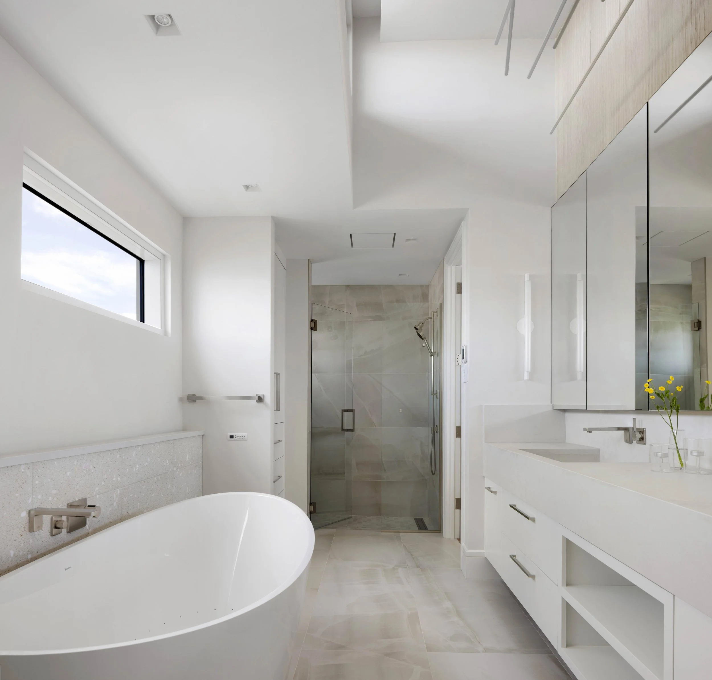Modern bathroom with white walls, a large window, and a walk-in shower with a glass door. There is a white bathtub, bathroom fixtures, and a minimalistic design with built-in cabinets and a small vase with yellow flowers.