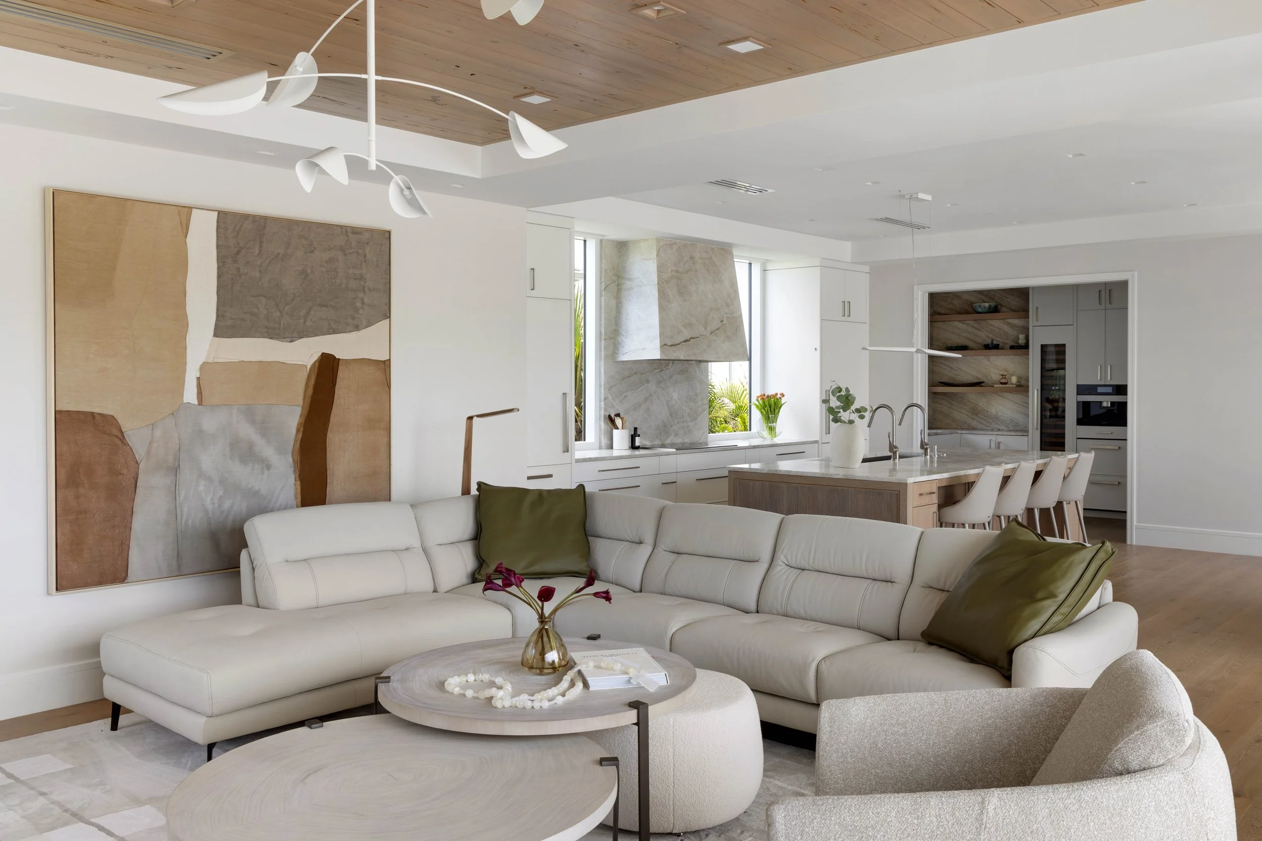 Modern living room with white curved sofa with green and beige pillows, abstract wall art, and a round coffee table with decor. Open kitchen in the background with built-in oven and wine fridge, wooden cabinets, and window with view of greenery.