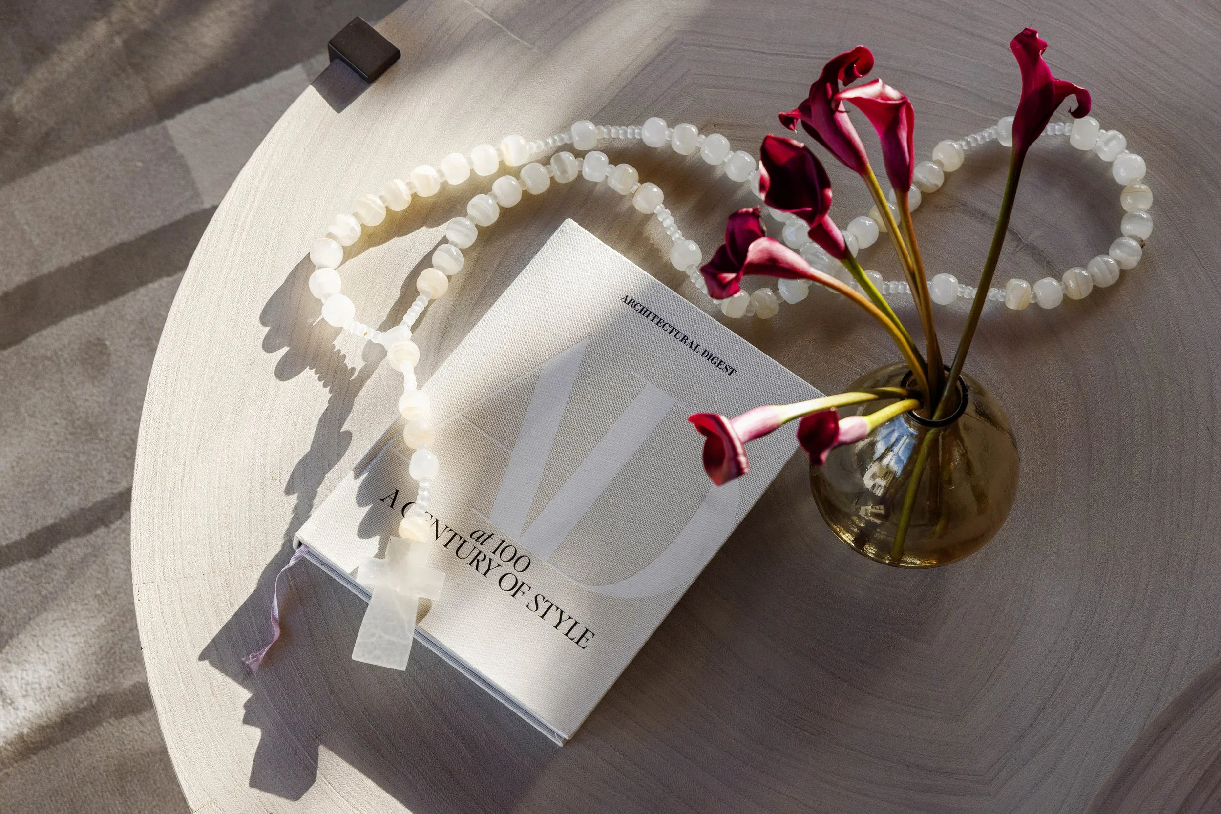 A white beaded necklace and a white gift box on a white surface with a booklet titled 'Architectural Digest' and a brown vase with dark pink calla lilies. Sunlight creates shadows on the surface.