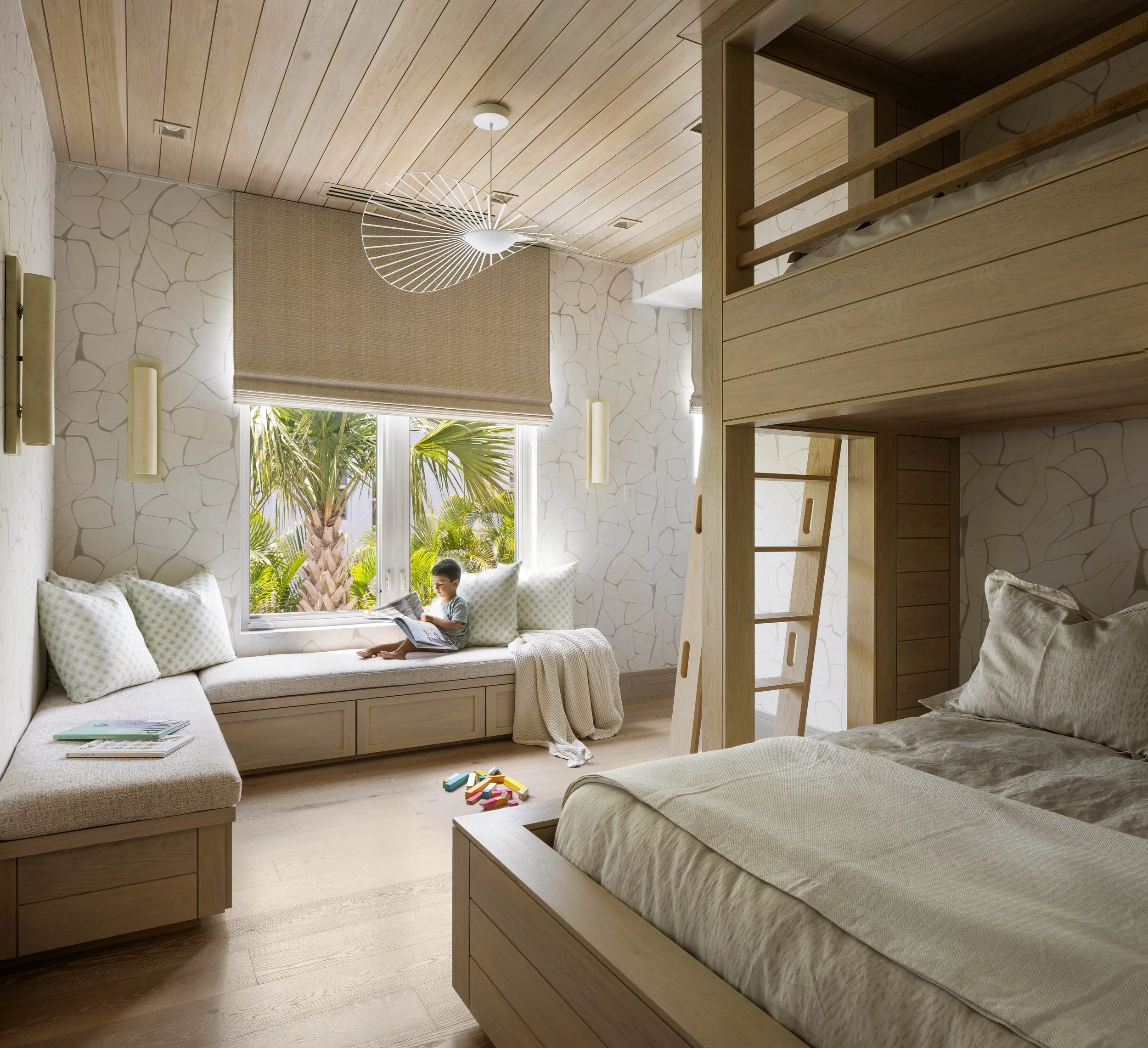 A cozy bedroom with light wood furniture and decor, a window with a view of tropical plants and palm trees, children playing with building blocks on the floor.
