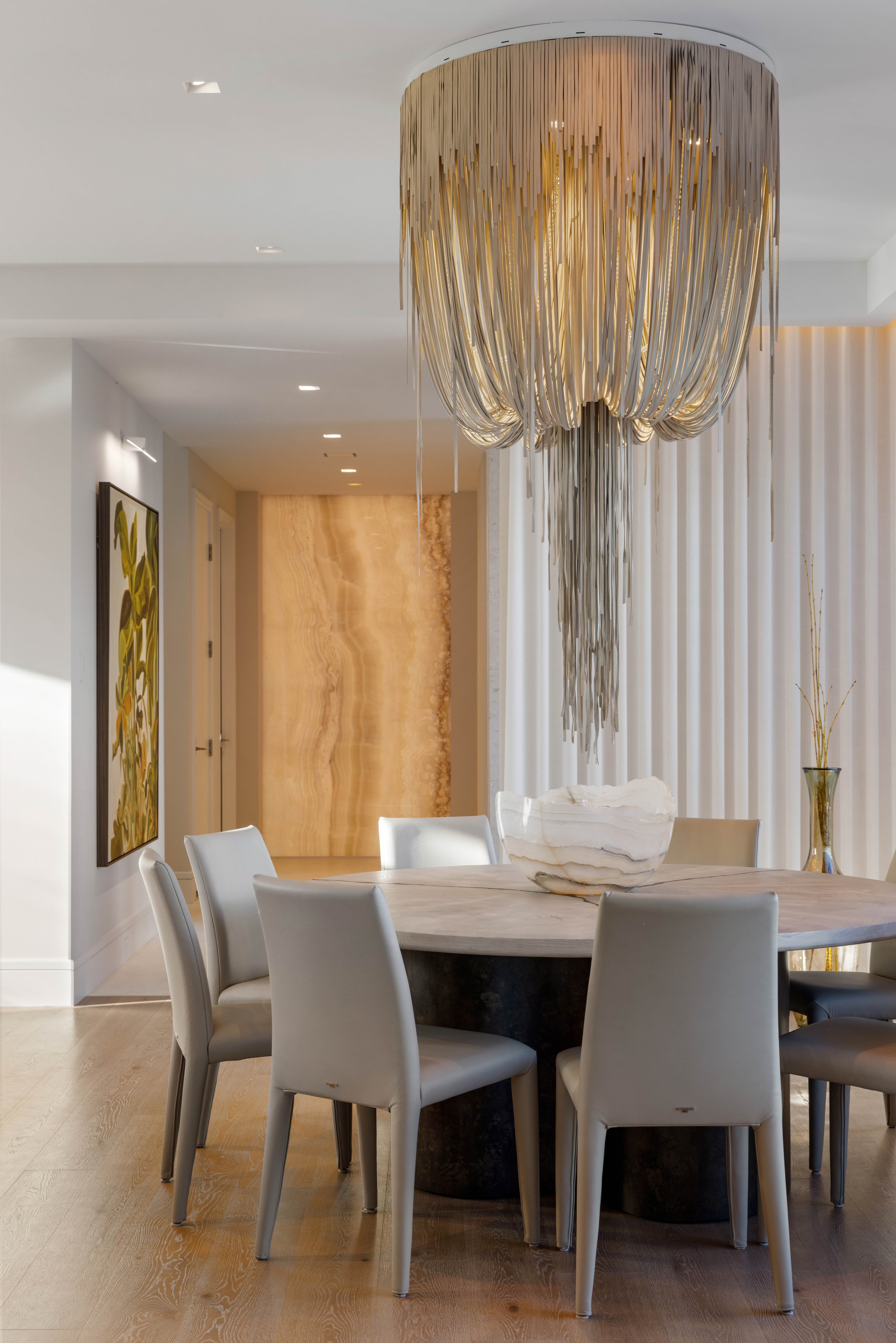 A round dining table with six white chairs in a modern, elegant room. A large marble bowl is on the table, with a decorative gold vase on the right side. A chandelier with gold and silver long strips hangs from the ceiling, illuminating the space. The background features white curtains and a marble wall with warm golden tones.