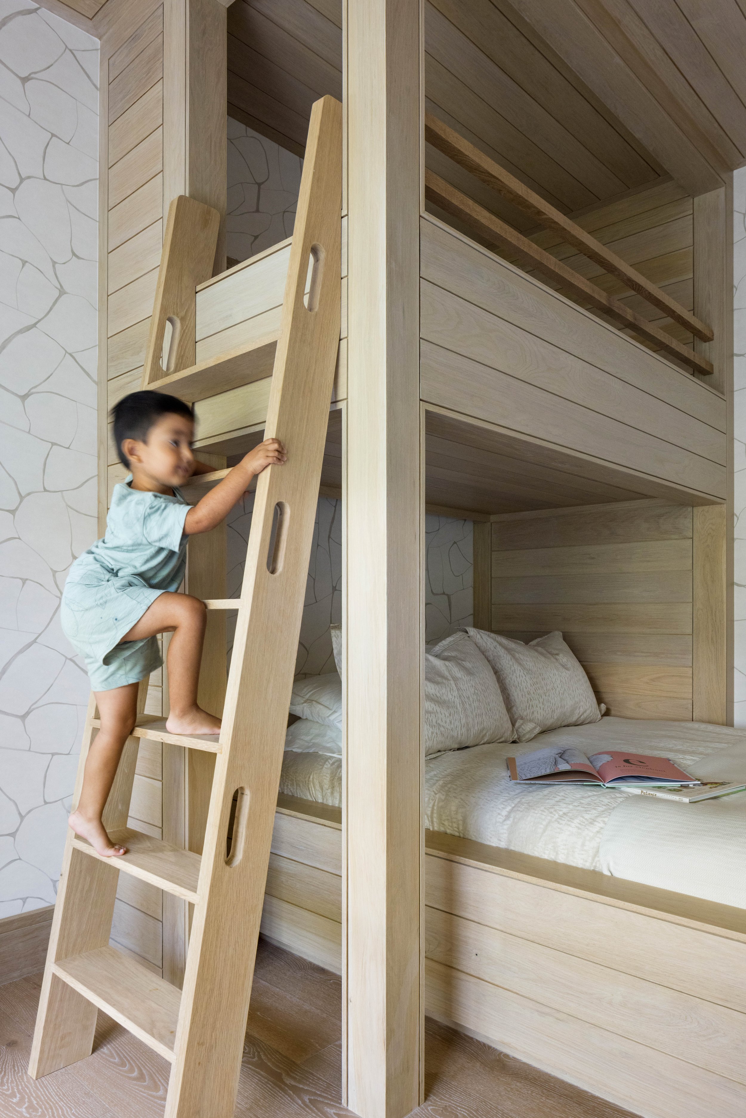 A young child climbing a wooden ladder leading to a bunk bed with a sleeping area and open books on the bed.