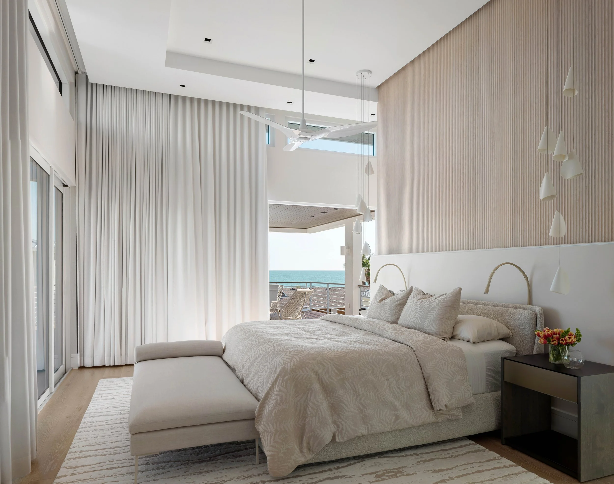 A modern bedroom with white walls, a beige upholstered bed with matching bedding, a black nightstand with a vase of flowers, a white ceiling fan, and large windows with white curtains overlooking a balcony and the ocean.