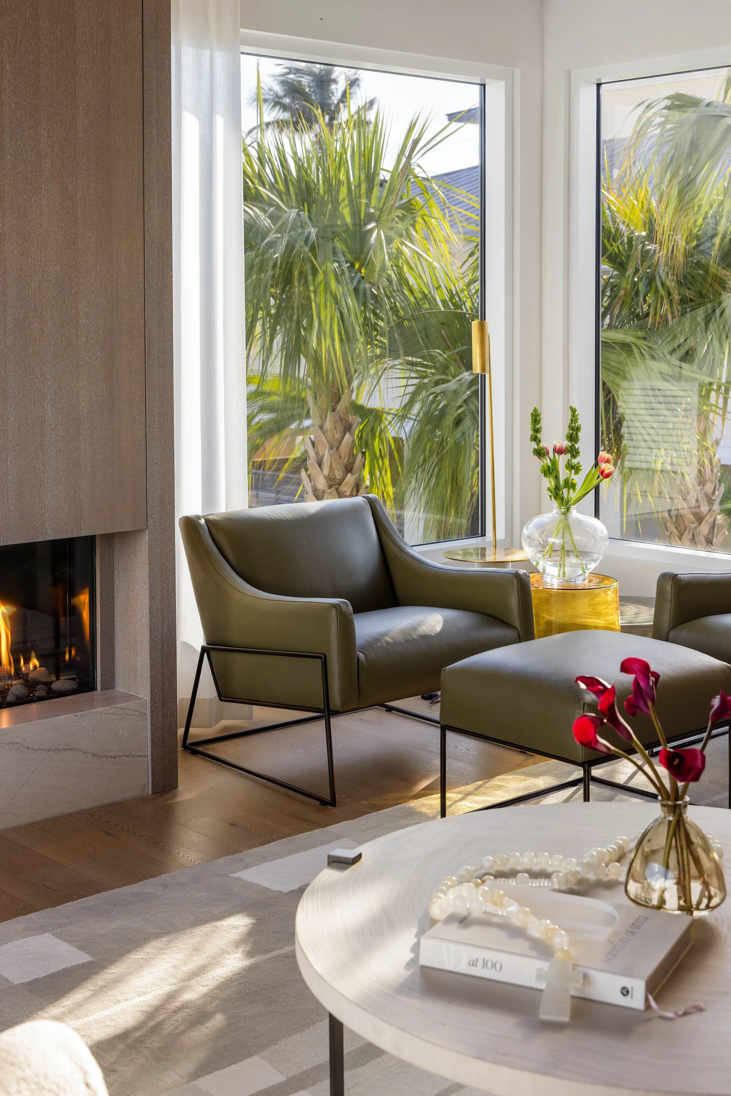 Living room with a large window showing green palm trees outside. There is a gray armchair, a side table with a gold lamp, a glass vase with pink and white tulips, and a white table with a pearl necklace and a book.