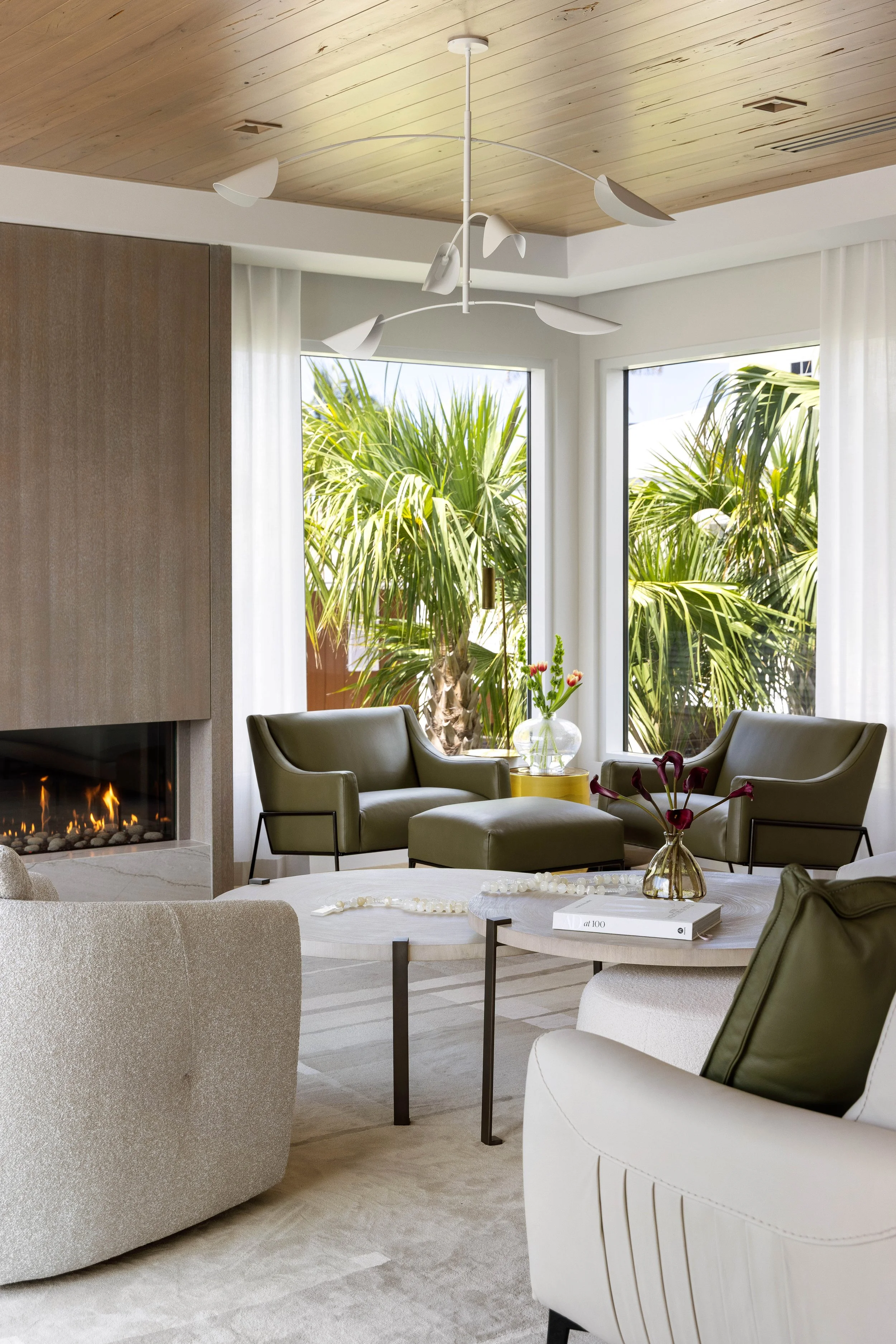 A modern living room with a wood-paneled ceiling, large windows, and outdoor greenery. There are green armchairs, a beige rounded armchair, a coffee table with books and vases with flowers, and a fireplace.