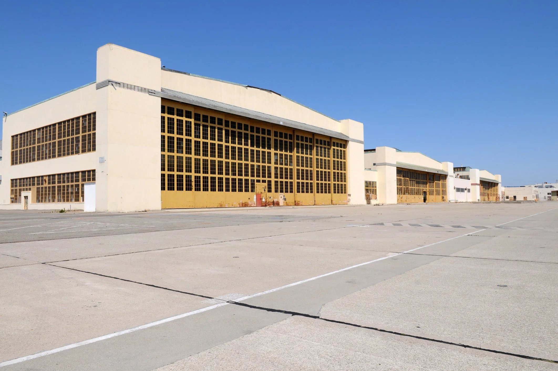 A large, empty parking lot in front of a beige industrial or warehouse building with a mostly clear blue sky overhead.