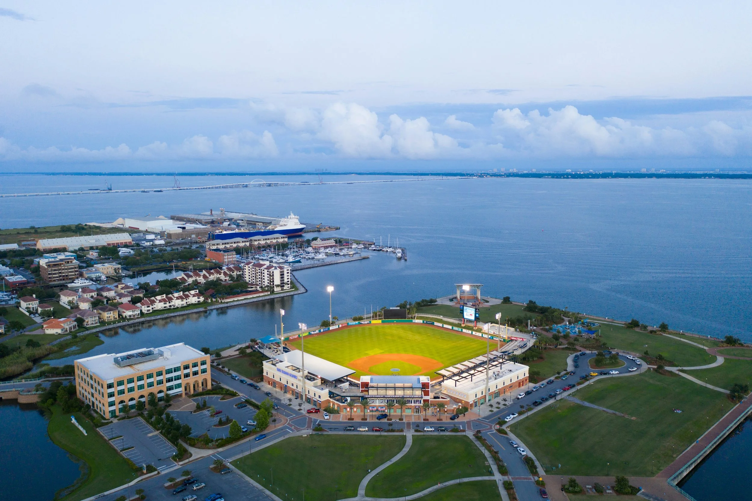 Aerial view of a coastal city with a marina, a baseball stadium, and a bridge over a river, with water and clouds in the background.
