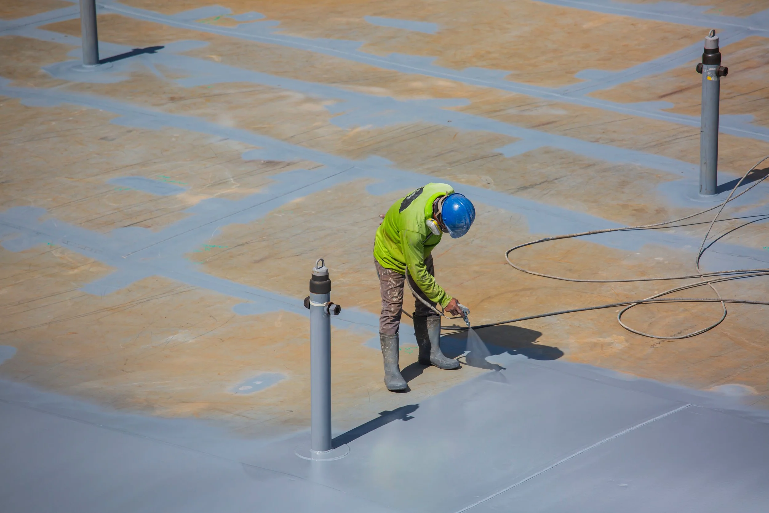 A construction worker wearing a blue safety helmet, neon green jacket, and gray gloves is applying a coating or sealant to a concrete surface on a building rooftop. The rooftop has several metal poles and a valve, with lines of sealing material.
