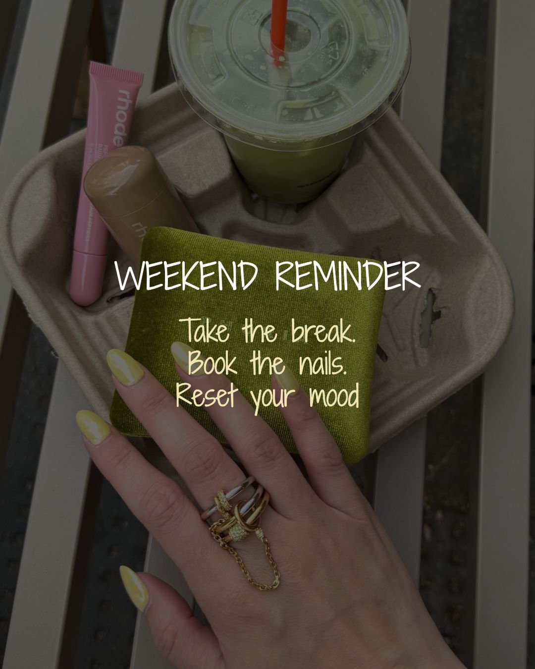 A hand with yellow painted nails holding a green cloth wallet over a beige drink holder containing a green smoothie with an orange straw, a pink tube, a small brown bottle, and a beige item on a white slatted surface, with overlaid text saying 'Weekend Reminder: Take the break. Book the nails. Reset your mood.'