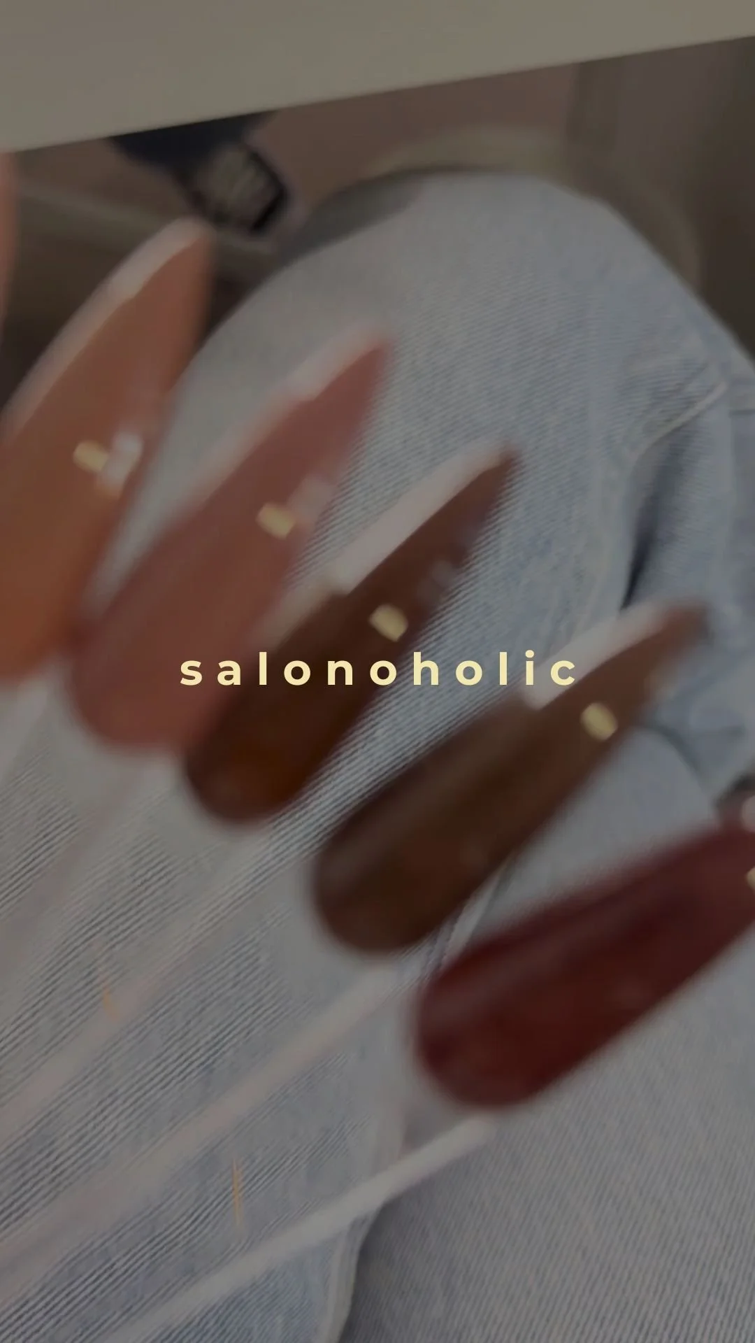 Close-up of a person's hand with long, square, nude-colored manicure resting on their knee, which is covered in light blue denim jeans. The background includes a silver laptop and a coffee cup, with the word "salonoholic" overlayed in beige font.
