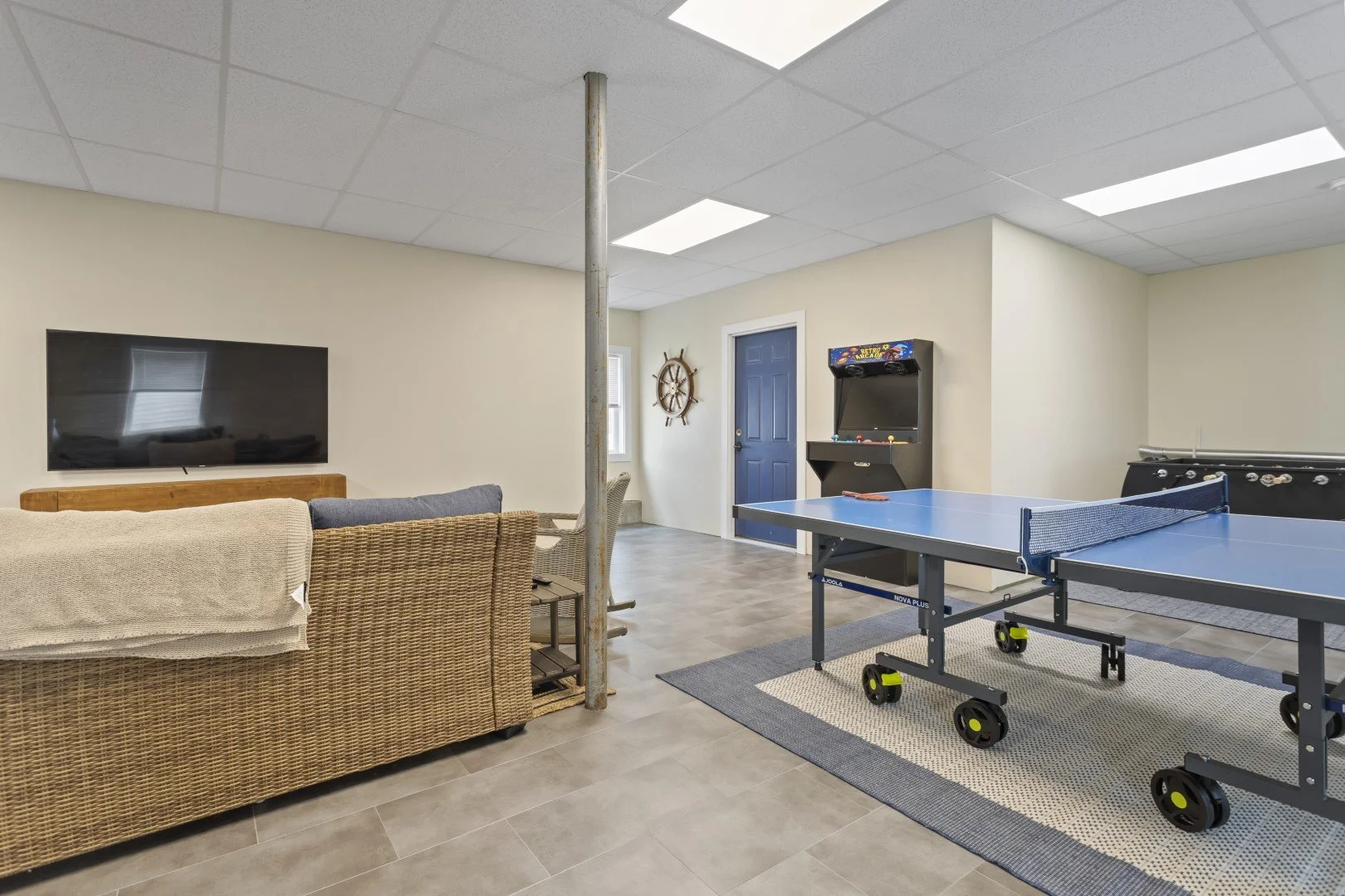 Recreational room with flat-screen TV, wicker sofa, foosball table, archery game, ping pong table, and an arcade game, with beige walls and gray tile flooring.
