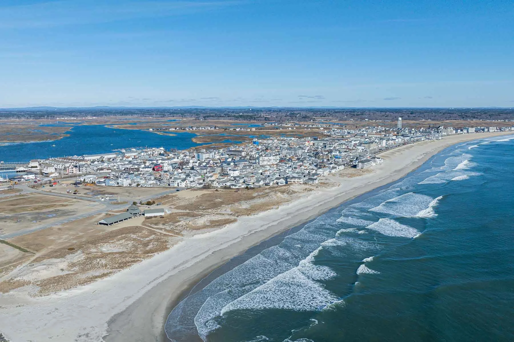An aerial view of a coastal town with a beach along the ocean, residential buildings, and a body of water in the background under a clear blue sky.