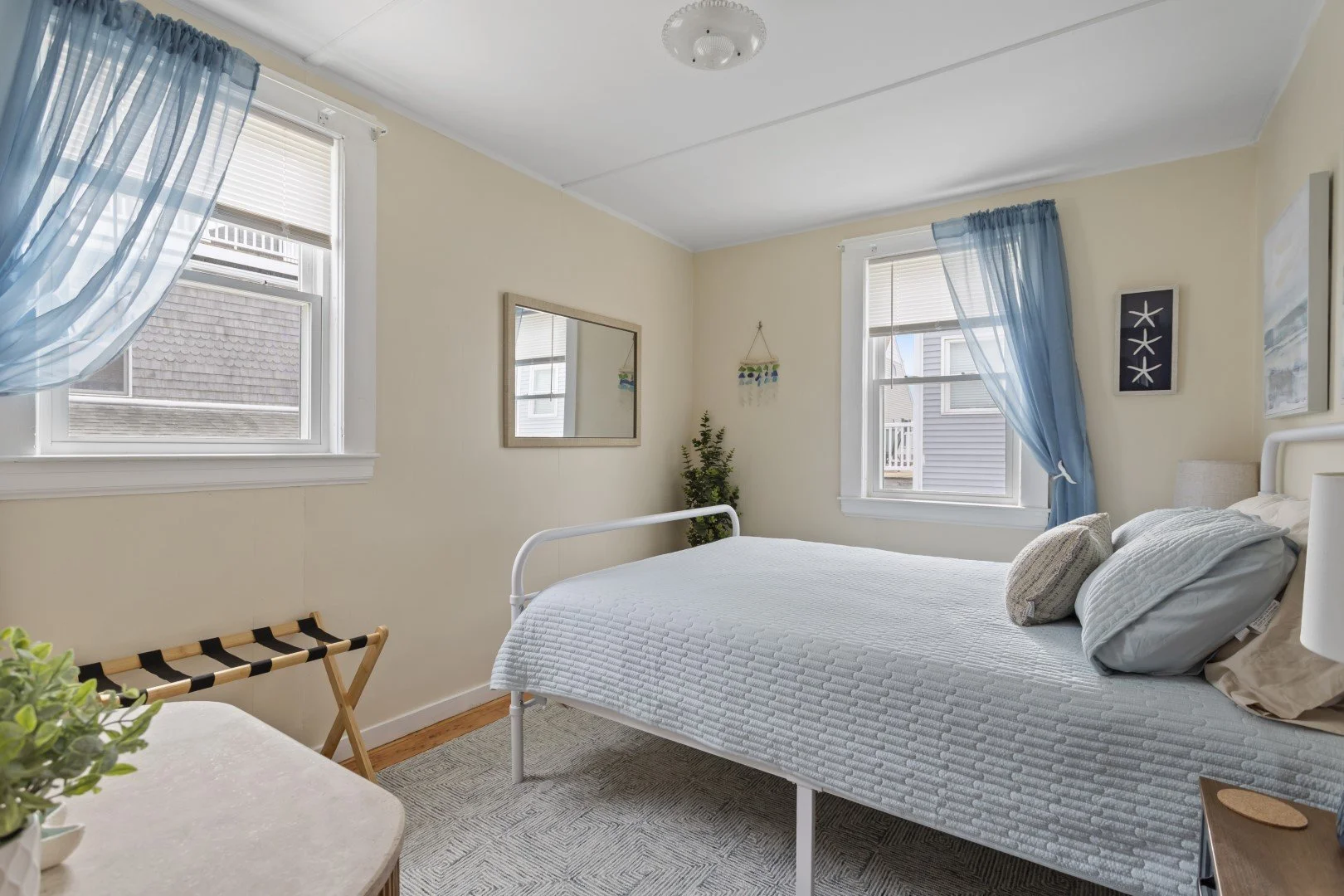 A bedroom with two windows covered by blue curtains, beige walls, a white bed with a textured quilt, and two pillows. There are picture frames and a mirror on the wall, a small plant, and a luggage rack near the window.