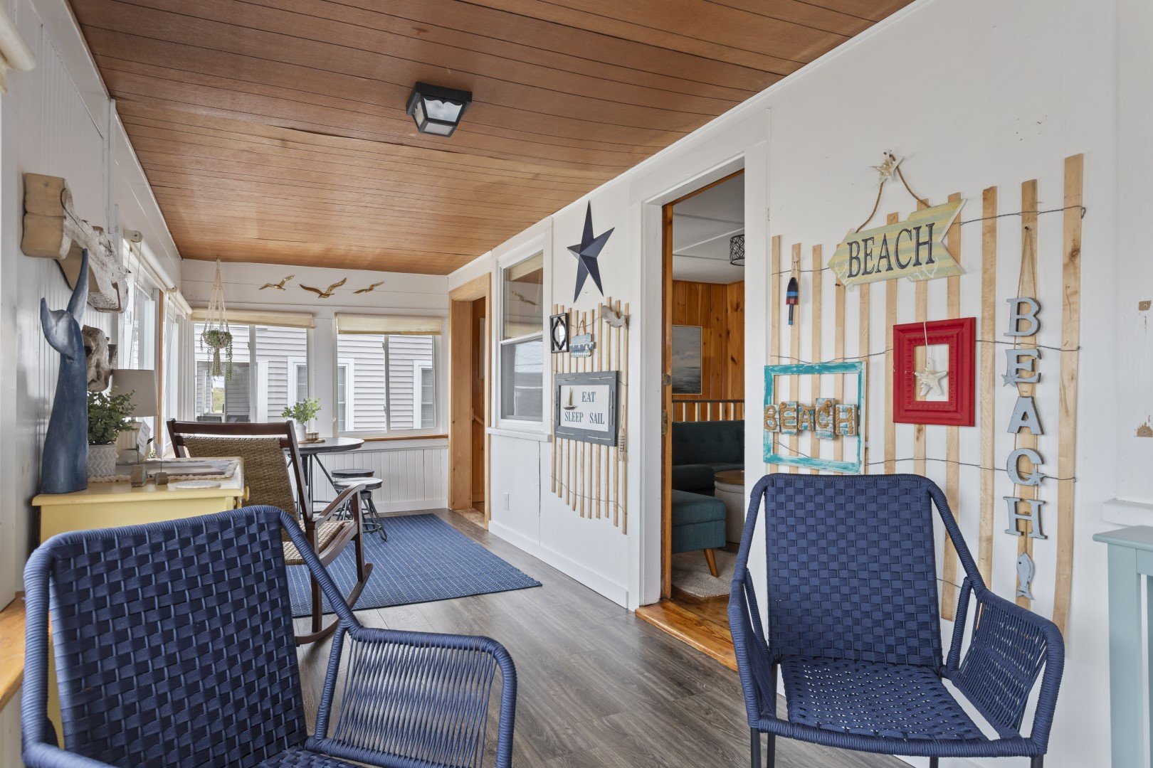 Coastal-themed sunroom with wooden ceiling, white walls, blue chairs, decorative beach signs, and ocean decor.