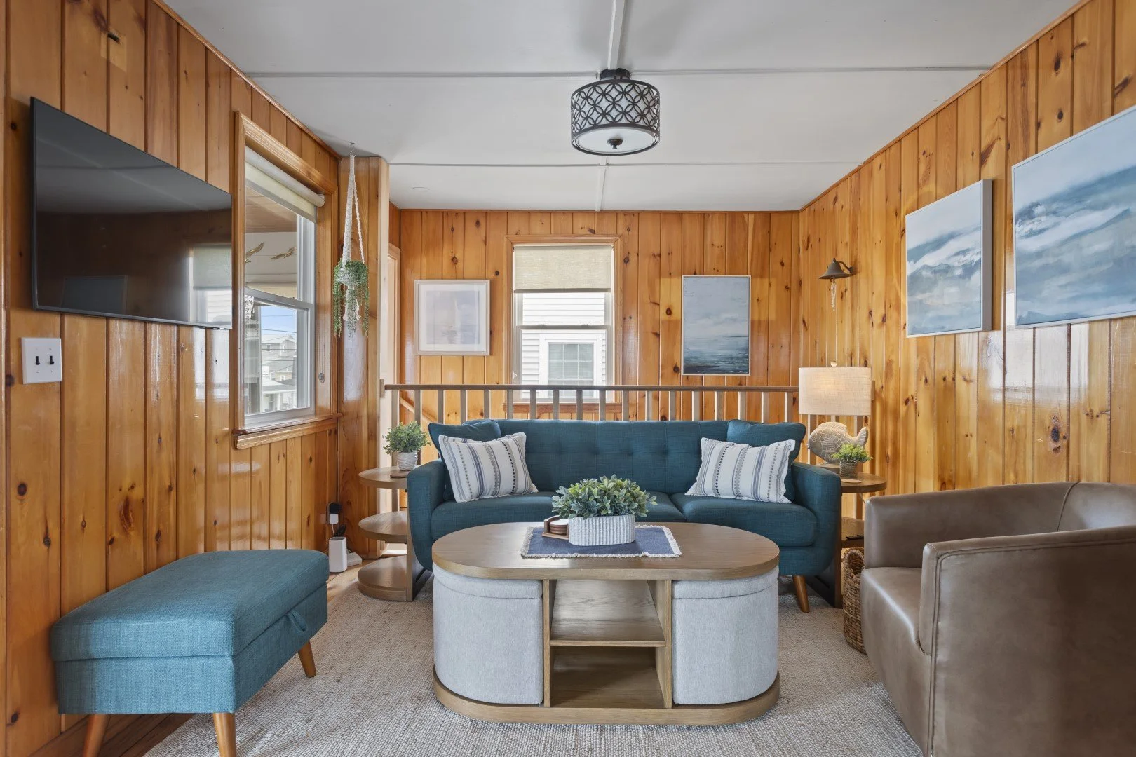 Living room with wood-paneled walls, a teal sofa with pillows, a wooden coffee table with plants, a beige armchair, a television on the wall, and artwork of seascapes, illuminated by a ceiling light and table lamp.