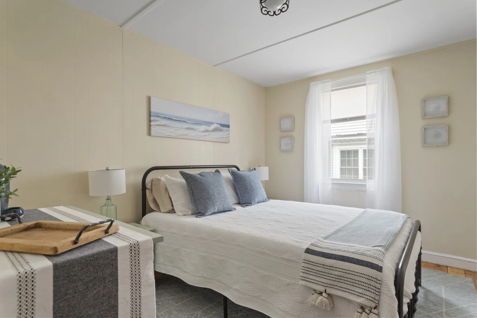 A bedroom with a bed, two blue pillows, and a framed ocean painting above the headboard. There are white curtains over a window, and a small side table with a lamp and tray beside the bed.