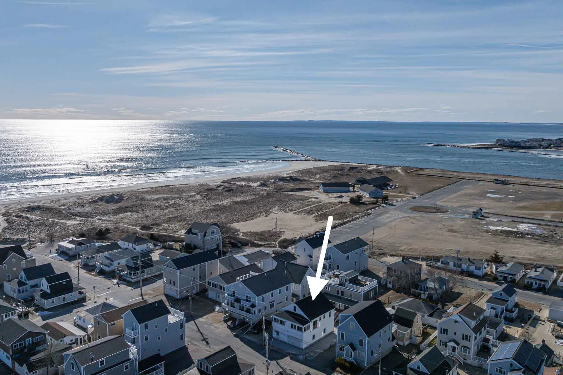 Aerial view of a coastal neighborhood with houses, a beach, water, and a breakwater with a pier extending into the ocean, under a partly cloudy sky.