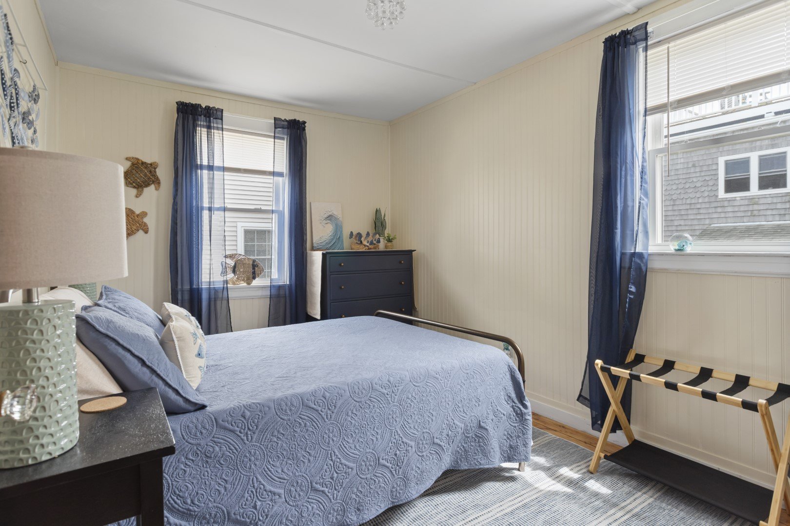 A cozy bedroom with a bed covered in a blue quilt, featuring pillows and decorative accents. The room has three windows with sheer navy curtains, natural light coming in, and ocean-themed decorations. A dark blue dresser with plants and artwork stands against the wall, and a luggage rack is near the window.
