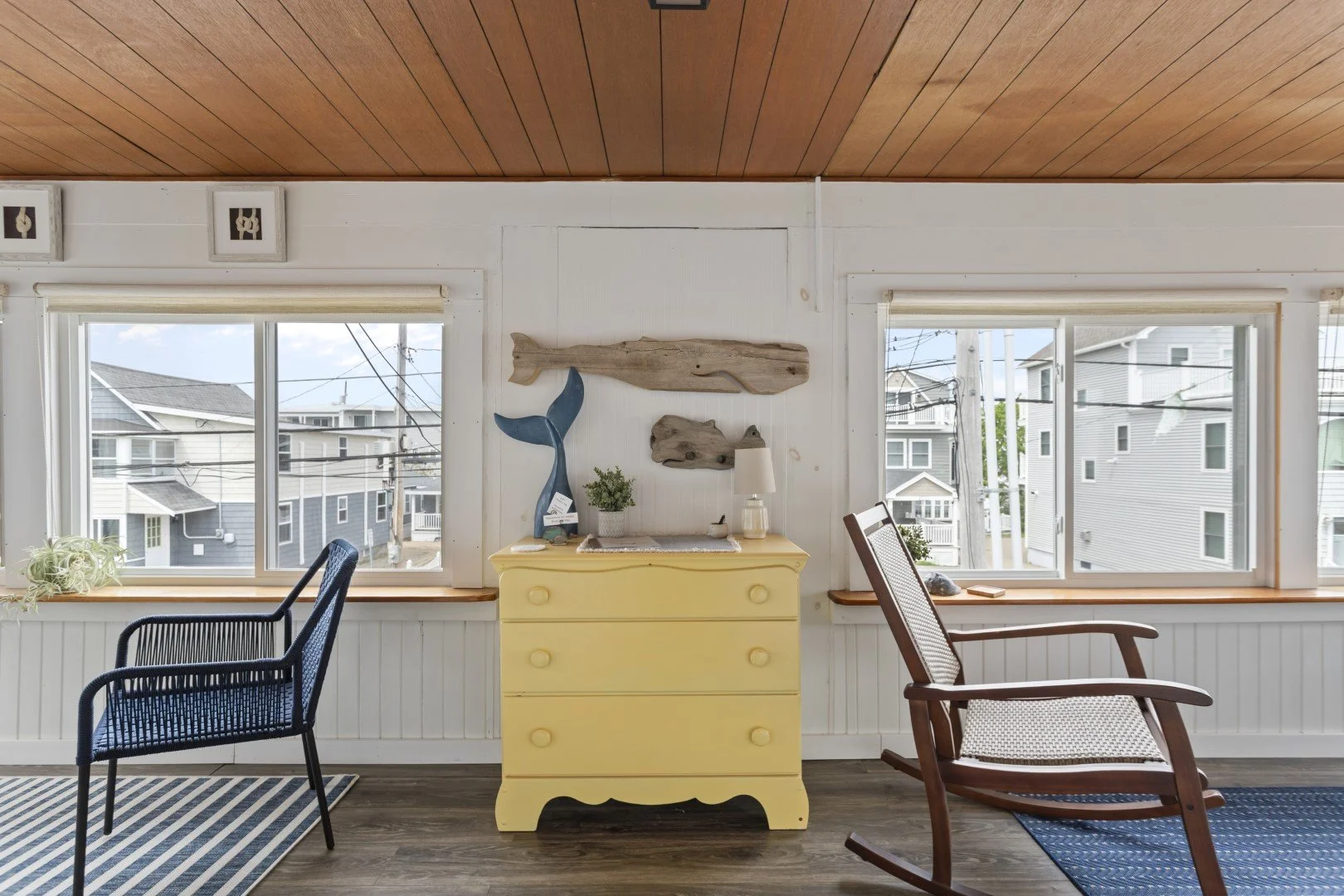 Interior of a room with two large windows showing neighboring houses, a yellow dresser with decor on top, a black chair on a striped rug, and a wooden rocking chair on a blue rug.