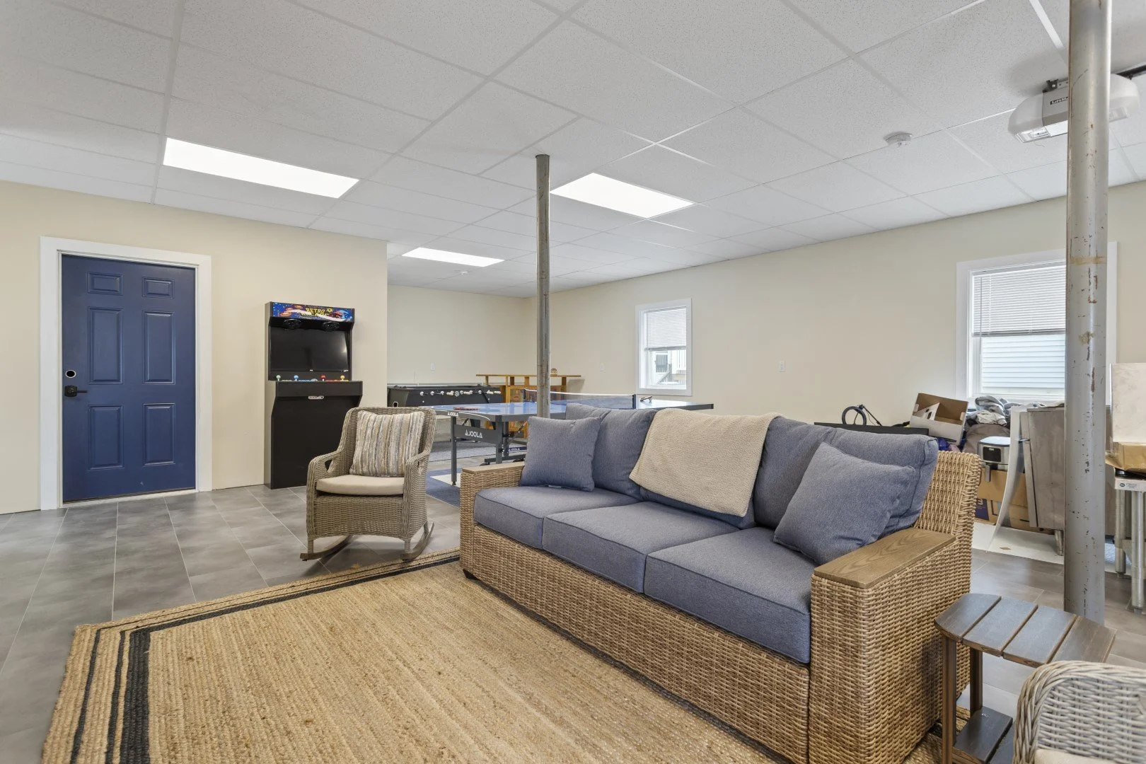 A recreational room with a wicker sofa with blue cushions, a wicker armchair, and a rug. There are two windows, a ping-pong table, a vintage arcade game, and boxes against the wall. A blue door is on the left.