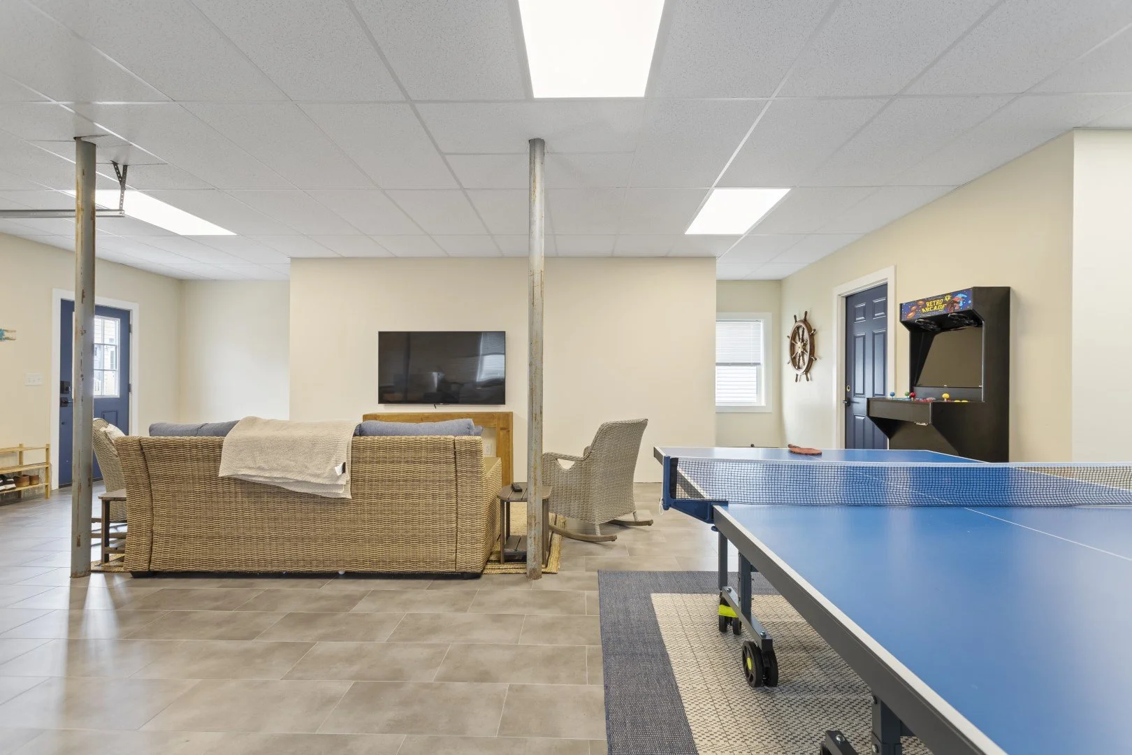 A recreational basement room with a ping pong table, a beige sofa, a television, a rock climbing wall, and arcade games, with a ceiling of white tiles and beige walls.