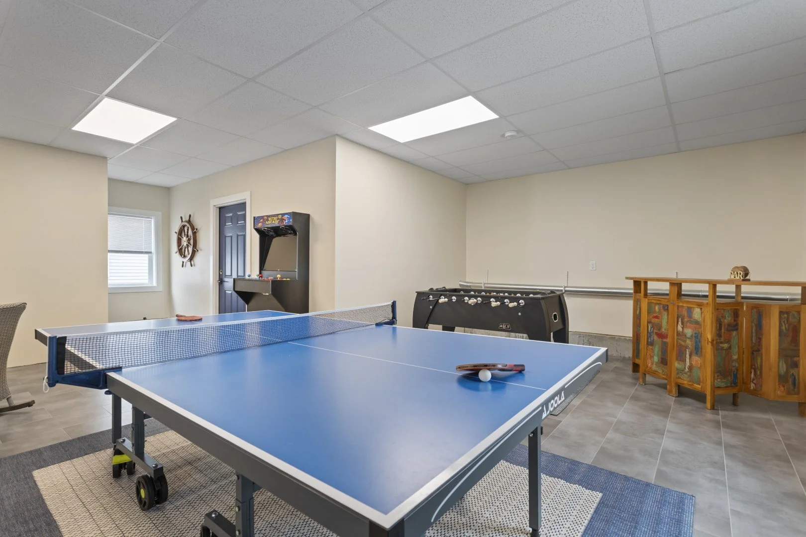 Indoor recreation room with blue ping pong table, ping pong paddles, and ball, arcade game, foosball table, and wooden bar or counter.