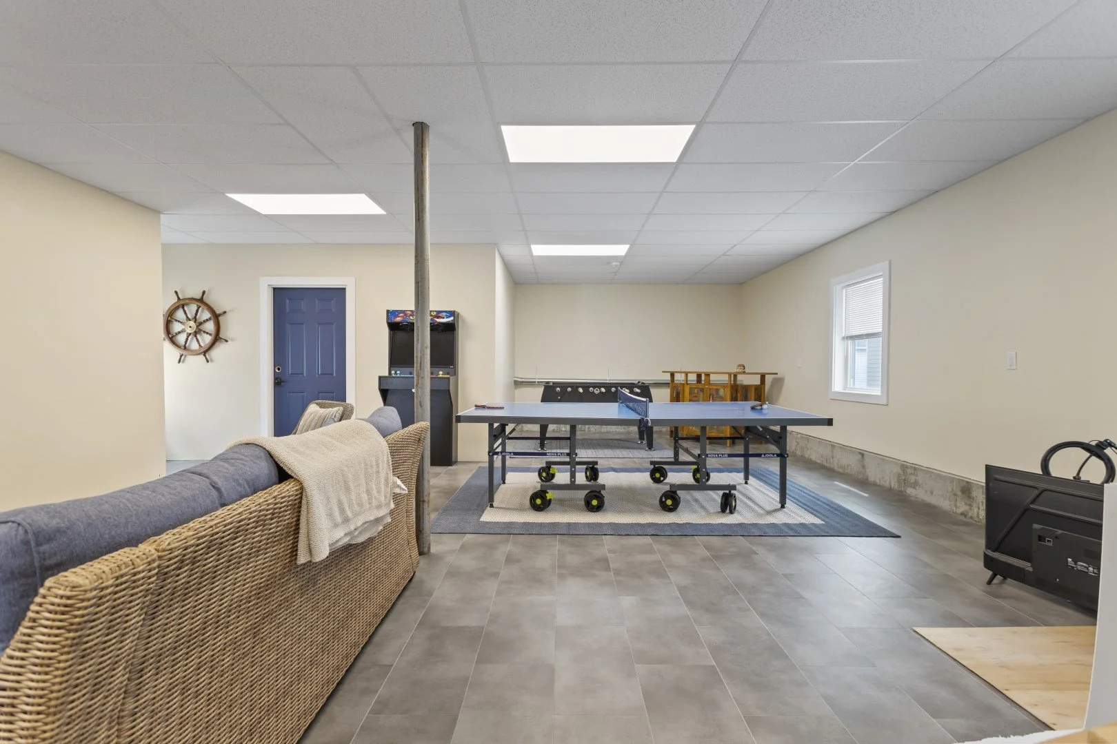 Basement recreation room with ping pong table, arcade games, foosball table, couch, window, and a blue door.