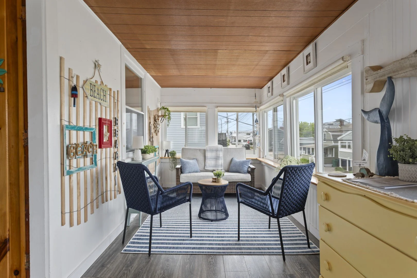 Cozy sunroom with white walls, wooden ceiling, blue chairs, striped rug, wicker loveseat with pillows, and large windows showing neighborhood houses and power lines.